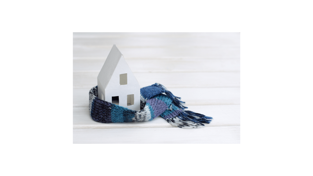 Image of a small white paper house sitting on a floor. There is a blue, green and black scarf wrapped around it to symbolize home insulation.