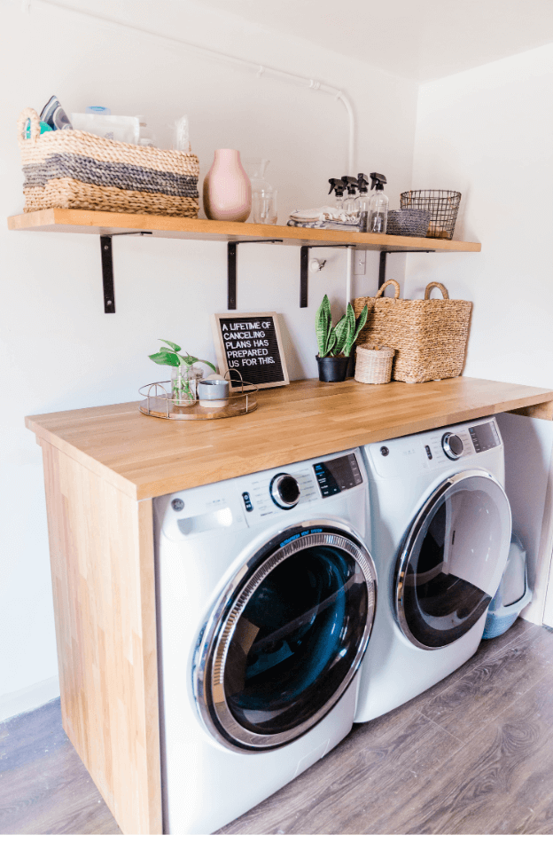 Image of a modern laundry room. Theres a washer and dryer with a slab of pretty wood going around it to create a table top on top. ABove that is a shelf. There are nicknacks around to complete the look.